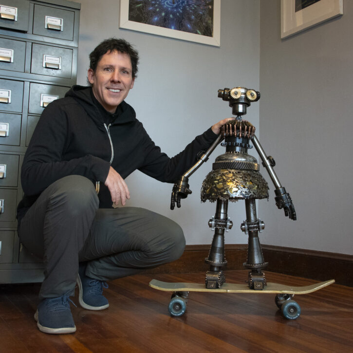 a sculptor kneels to pose next to a steel sculpture of a little girl in a steel skirt. She rests on top of a skateboard.